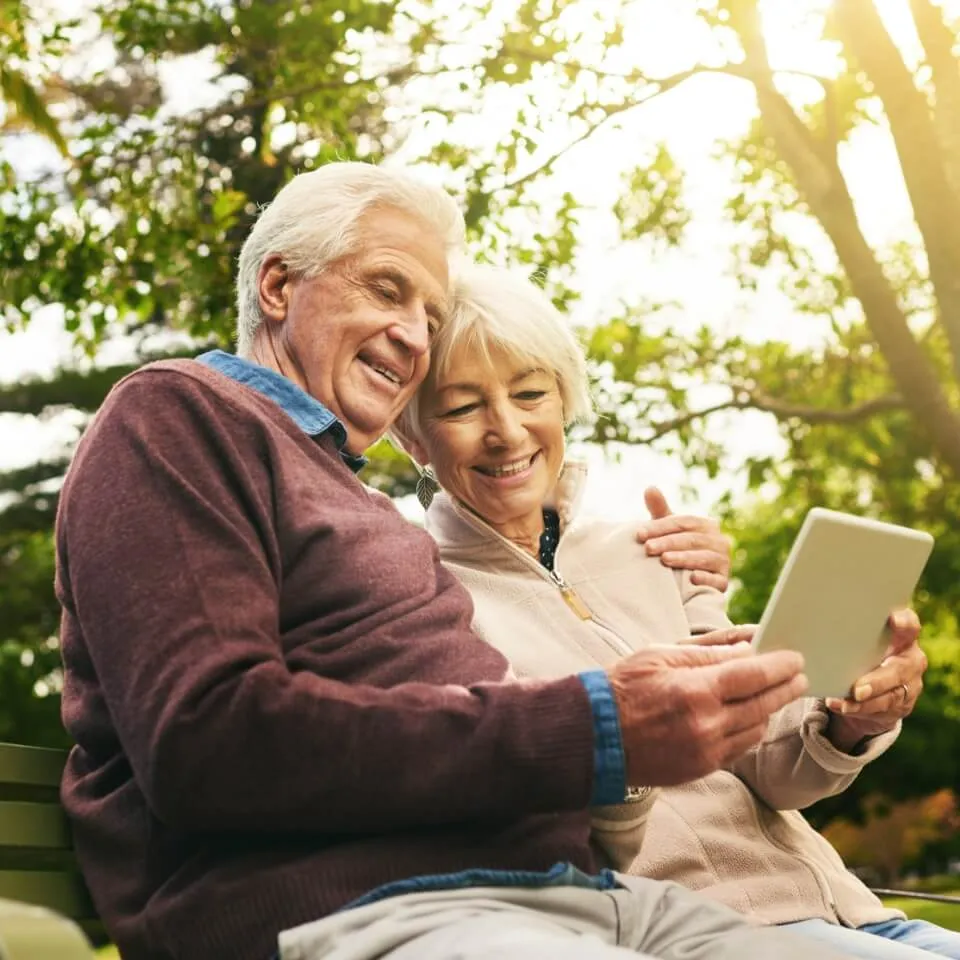 Elderly couple sitting on a park bench, smiling warmly as they share a moment looking at a tablet. Sunlight filters through trees, creating a joyful atmosphere.